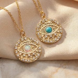 Puede incluir: Dos collares dorados con colgantes redondos con diseño de ojo. Uno tiene un centro turquesa, el otro un ópalo. Ambos están rodeados de pequeñas piedras transparentes. Descansan sobre una tela color crema.