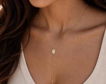 Puede incluir: Un collar tipo lariat en tono dorado con una cadena fina y pequeñas cuentas espaciadas uniformemente. El collar incluye un colgante ovalado central con una figura religiosa y una pequeña cruz que cuelga al final de la cadena vertical.