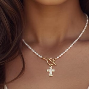 May include: A necklace featuring a strand of small white pearls connected to a gold-toned toggle clasp. A gold-toned cross pendant with a textured, layered design hangs from the center of the toggle clasp, resting against the collarbone area.