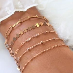 Pode incluir: Cinco pulseiras douradas com diferentes estilos de corrente, incluindo uma corrente com discos redondos, uma corrente com elos retangulares, uma corrente com pequenas contas redondas e uma corrente simples. As pulseiras são exibidas em um fundo branco.