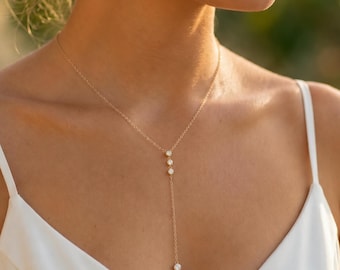May include: A delicate gold lariat necklace featuring three small, clear, round-cut stones set in a vertical line, suspended from a thin gold chain. The necklace is worn with a white top and a small, single, clear stone stud earring.