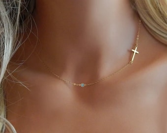 May include: A delicate gold chain necklace with a small blue gemstone and a sideways cross pendant.