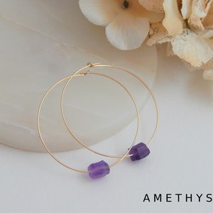 May include: Gold-tone hoop earrings with raw amethyst gemstones. The earrings are round and feature a small, rough-cut purple amethyst stone at the bottom. The word "AMETHYST" is printed in black, sans-serif font at the bottom of the image.