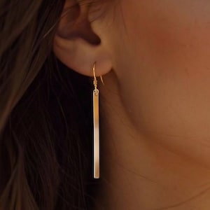 May include: A close-up shot of a person wearing gold bar earrings. The earrings are long and rectangular, hanging from the ear. The person has brown hair and is looking towards the right side of the frame. The background is blurred.