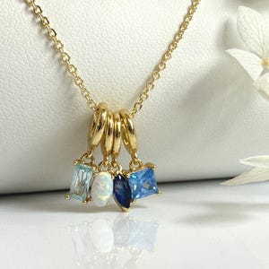 May include: A gold-colored necklace with a delicate chain. The necklace features four charms: a rectangular light blue stone, an oval opal, a dark blue teardrop, and a square light blue stone. The charms hang from a set of gold rings.