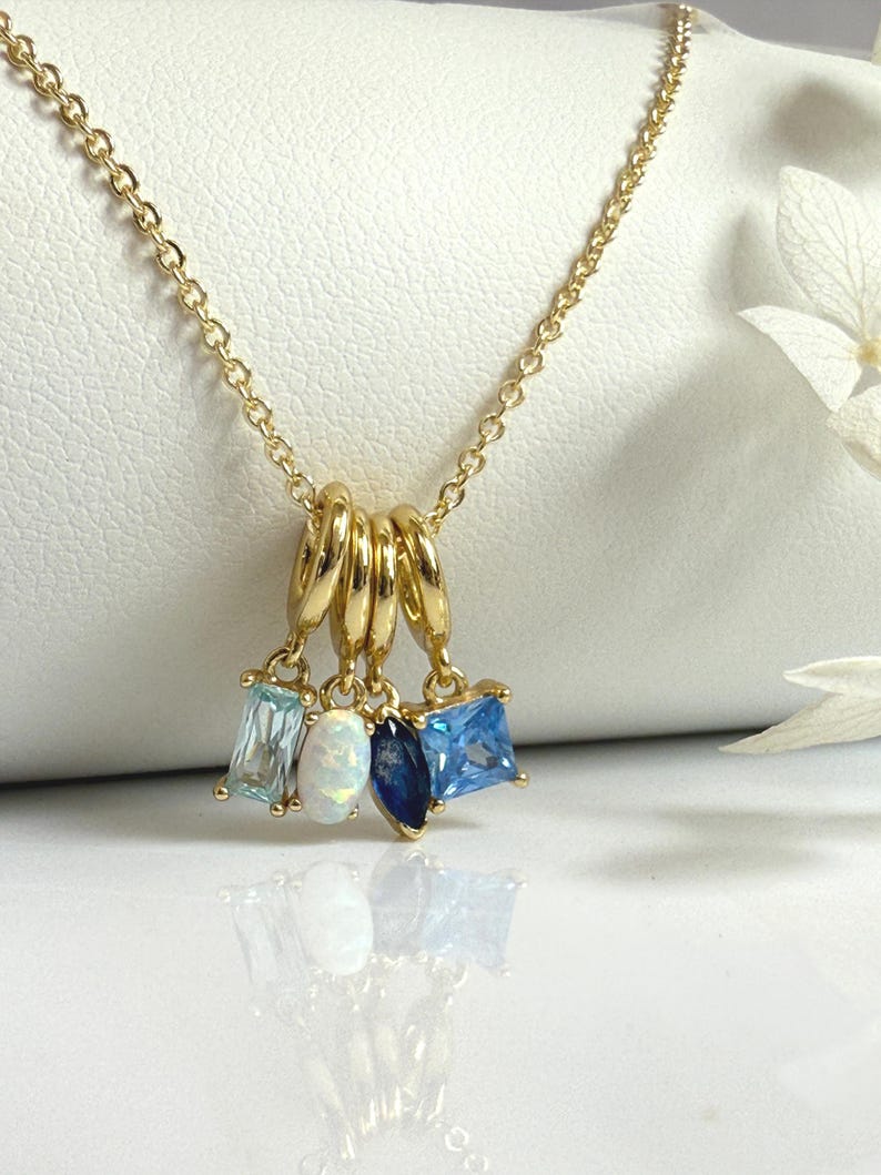 May include: A gold-colored necklace with a delicate chain. The necklace features four charms: a rectangular light blue stone, an oval opal, a dark blue teardrop, and a square light blue stone. The charms hang from a set of gold rings.
