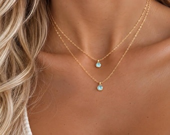 May include: A layered gold chain necklace featuring two delicate strands. Each strand holds a small, round, light blue gemstone pendant set in a gold bezel. The chains are thin and metallic, resting against the neck with a polished finish.