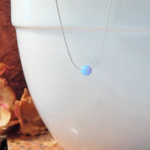 May include: A delicate silver chain necklace with a single, round, iridescent opal pendant. The opal is a light blue colour with hints of pink and white.