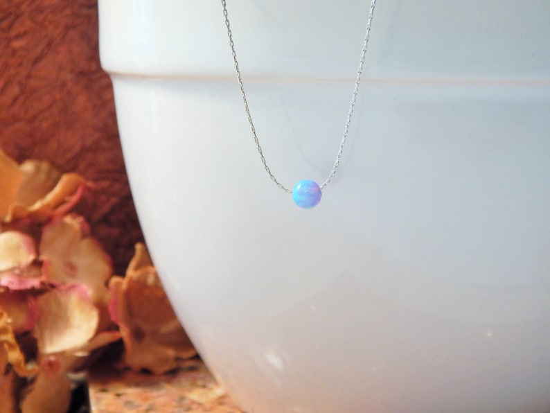 May include: A delicate silver chain necklace with a single, round, iridescent opal pendant. The opal is a light blue colour with hints of pink and white.
