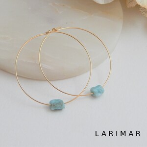 May include: Gold-tone hoop earrings featuring a raw, light blue Larimar stone. The earrings have a simple, elegant design with a minimalist aesthetic. The word "LARIMAR" is printed in black at the bottom of the image.