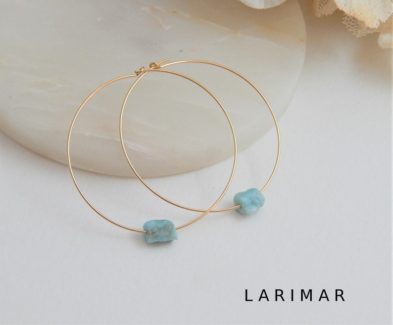 May include: Gold-tone hoop earrings featuring a raw, light blue Larimar stone. The earrings have a simple, elegant design with a minimalist aesthetic. The word "LARIMAR" is printed in black at the bottom of the image.