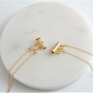 May include: Two gold-colored necklace clasps with delicate chain attachments. The clasps are cylindrical with a T-bar closure. The chains are fine and dangle from the clasps, set against a white, circular background.