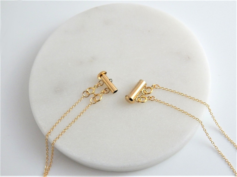 May include: Two gold-colored necklace clasps with delicate chain attachments. The clasps are cylindrical with a T-bar closure. The chains are fine and dangle from the clasps, set against a white, circular background.