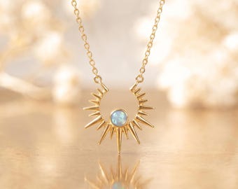 May include: A gold-toned necklace featuring a sunburst-shaped pendant with a central, round, iridescent blue-white stone. The pendant is suspended from a delicate gold-toned chain, with the design radiating outward in pointed metal rays.