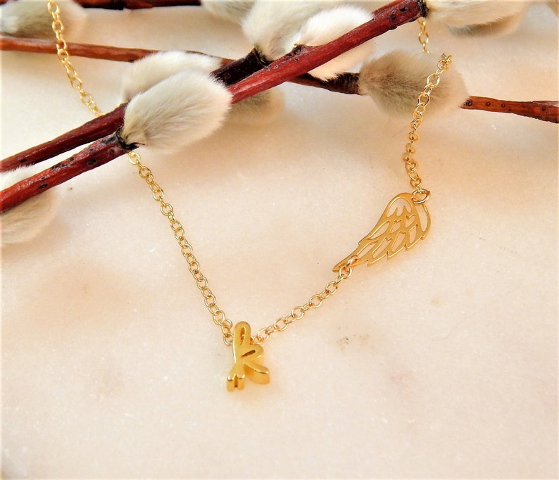 Puede incluir: Un collar de cadena dorada con un peque&ntilde;o dije de ala dorada y un dije de letra K dorada.