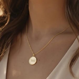 May include: A gold-colored necklace with a delicate chain and a circular pendant. The pendant features the number "1111" in a simple font. The necklace is worn against a light background.
