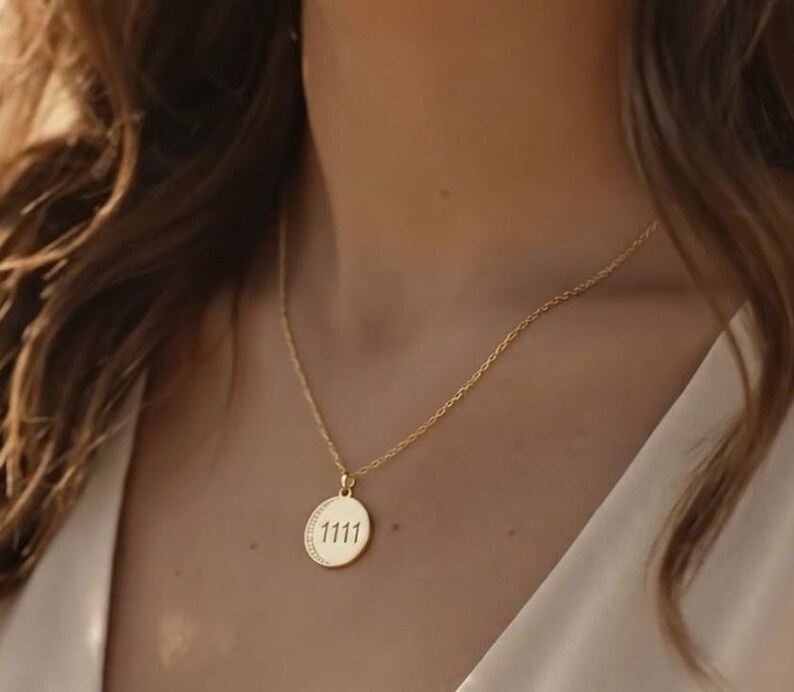 May include: A gold-colored necklace with a delicate chain and a circular pendant. The pendant features the number "1111" in a simple font. The necklace is worn against a light background.