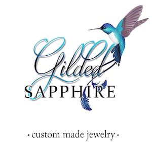May include: A logo featuring a blue and white hummingbird in flight above the text Gilded SAPPHIRE in a stylized script and serif font, with the words custom made jewelry positioned below the main text, all set against a plain white background.