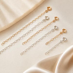 May include: A collection of six jewelry extender chains in gold and silver tones, each featuring a delicate oval link design and a circular spring ring clasp at one end, arranged in a diagonal pattern on a neutral textured surface.