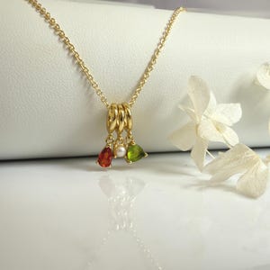 May include: A gold-colored necklace with a pendant featuring three rings and three teardrop-shaped gemstones in red, white, and green. The necklace is displayed on a white surface with white flowers in the background.
