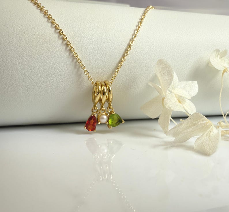May include: A gold-colored necklace with a pendant featuring three rings and three teardrop-shaped gemstones in red, white, and green. The necklace is displayed on a white surface with white flowers in the background.