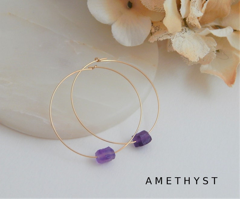 May include: Gold-tone hoop earrings with raw amethyst gemstones. The earrings are round and feature a small, rough-cut purple amethyst stone at the bottom. The word "AMETHYST" is printed in black, sans-serif font at the bottom of the image.