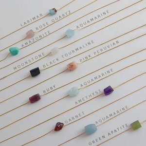 May include: A collection of delicate necklaces featuring various gemstones, including larimar, rose quartz, and aquamarine. Each necklace has a gold chain and a single, colorful stone. The names of the gemstones are printed above each necklace.