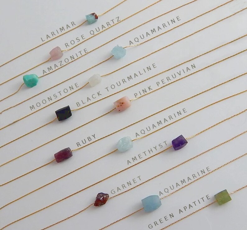 May include: A collection of delicate necklaces featuring various gemstones, including larimar, rose quartz, and aquamarine. Each necklace has a gold chain and a single, colorful stone. The names of the gemstones are printed above each necklace.