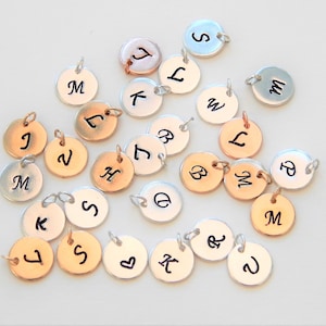 May include: A collection of small, round metal charms with individual letters engraved in black. The charms are in various metallic finishes, including silver, gold, and rose gold. Each charm has a small loop for attaching to a chain or bracelet.