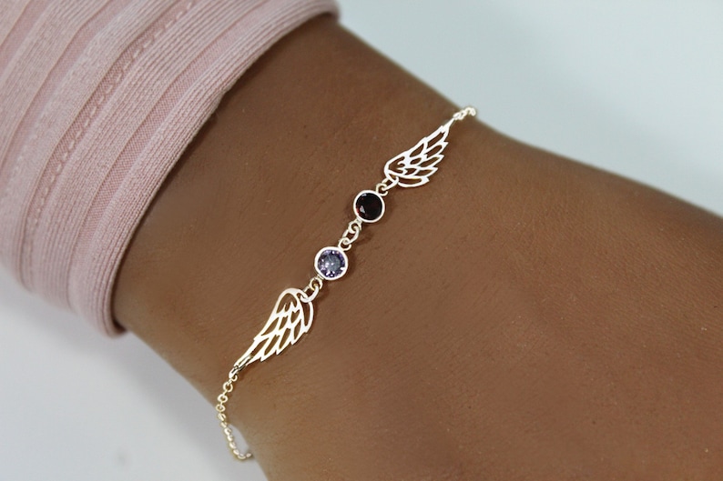 K&ouml;nnte beinhalten: Ein goldenes Kettenarmband mit zwei zarten Engelsfl&uuml;gel-Anh&auml;ngern und drei kleinen Edelsteinen. Die Edelsteine sind lila, rot und lila.