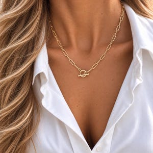 May include: A gold-toned necklace featuring elongated rectangular chain links and a circular toggle clasp closure. The necklace rests against a white collared shirt, showcasing a polished metallic finish and a structured, uniform link design throughout.