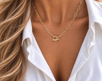 May include: A gold-toned necklace featuring elongated rectangular chain links and a circular toggle clasp closure. The necklace rests against a white collared shirt, showcasing a polished metallic finish and a structured, uniform link design throughout.