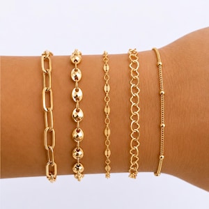 May include: Five gold-coloured bracelets of varying designs are displayed on a wrist. The bracelets include a chain link, a series of oval links, a delicate chain, a heart-shaped link, and a beaded chain. The bracelets are made of a shiny, gold-coloured metal.