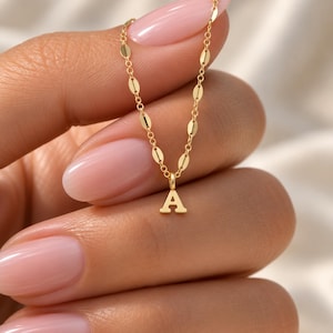 May include: A gold-toned necklace featuring a delicate chain with small, flat, oval-shaped links and a single, centered pendant in the shape of the capital letter A. The metal has a polished, reflective finish, creating a bright, metallic appearance.