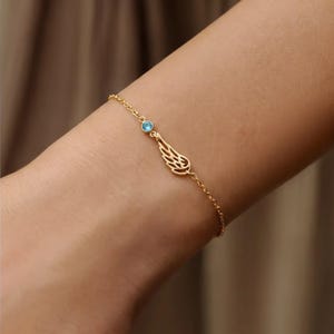May include: A delicate gold-colored bracelet featuring a small, light blue gemstone and a stylized wing charm. The bracelet is worn on a wrist, showcasing its fine chain and intricate design. The wing charm is a detailed cutout.