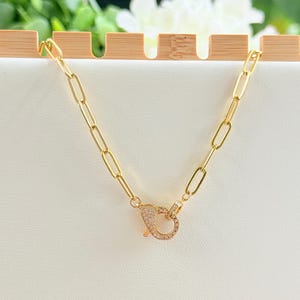 May include: A gold-colored chain necklace with an oval link design. The necklace features a decorative clasp encrusted with small, clear crystals. The jewelry is displayed on a white surface with a wooden accent in the background.