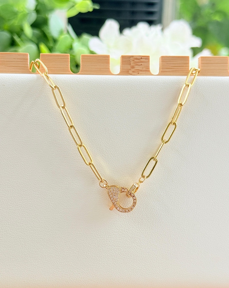 May include: A gold-colored chain necklace with an oval link design. The necklace features a decorative clasp encrusted with small, clear crystals. The jewelry is displayed on a white surface with a wooden accent in the background.