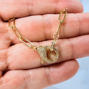 May include: A gold-colored necklace clasp with a rectangular link chain. The clasp is encrusted with small, clear gemstones. The jewelry is displayed against a skin-toned background.