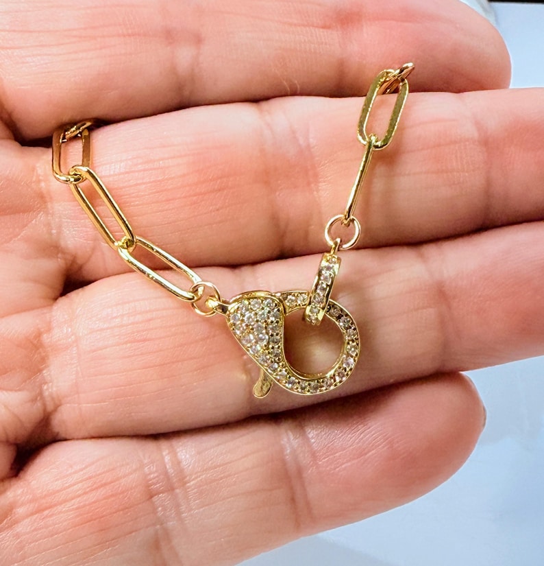 May include: A gold-colored necklace clasp with a rectangular link chain. The clasp is encrusted with small, clear gemstones. The jewelry is displayed against a skin-toned background.