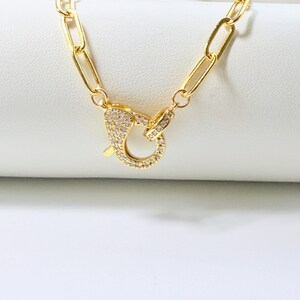 May include: Gold-colored necklace with a large, ornate clasp. The clasp is encrusted with small, clear gemstones. The chain links are elongated and rectangular. The jewelry is displayed on a white surface.