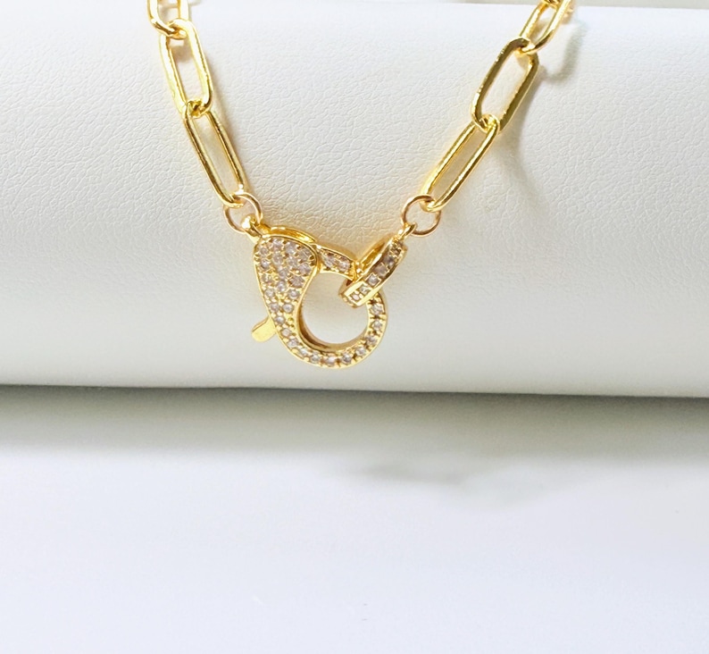 May include: Gold-colored necklace with a large, ornate clasp. The clasp is encrusted with small, clear gemstones. The chain links are elongated and rectangular. The jewelry is displayed on a white surface.