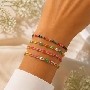 Puede incluir: Cuatro pulseras de cuentas de colores se muestran en una muñeca. Las pulseras presentan pequeñas cuentas redondas en varios colores, incluyendo rojo, naranja, amarillo, verde y azul. Las pulseras están conectadas por una cadena dorada.