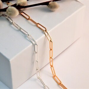 May include: Two paperclip chains, one in a polished silver tone and one in a polished gold tone, draped vertically. The chains feature elongated, rectangular links connected in a repeating pattern, showcasing a metallic finish and smooth texture.