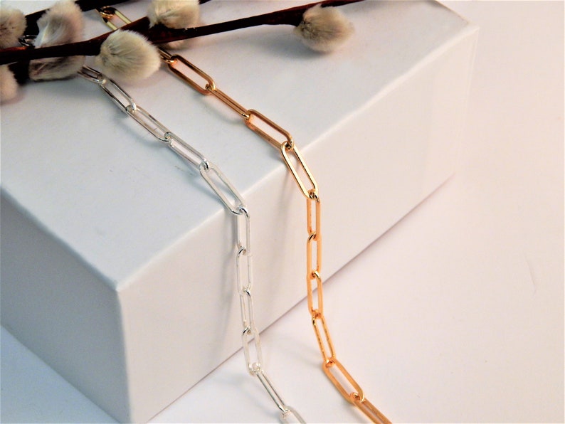 May include: Two paperclip chains, one in a polished silver tone and one in a polished gold tone, draped vertically. The chains feature elongated, rectangular links connected in a repeating pattern, showcasing a metallic finish and smooth texture.