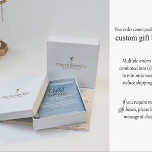 K&ouml;nnte beinhalten: Wei&szlig;e Geschenkbox mit einem Kolibri-Logo und dem Text "Gilded Sapphire" auf der Oberseite. Die Box ist ge&ouml;ffnet und eine kleinere wei&szlig;e Box mit einer blauen Karte darin ist sichtbar. Die Karte tr&auml;gt den Text "Gilded Sapphire". Der Text "Ihre Bestellung wird in unseren individuellen Geschenkboxen verpackt." ist auf der rechten Seite des Bildes zu sehen.