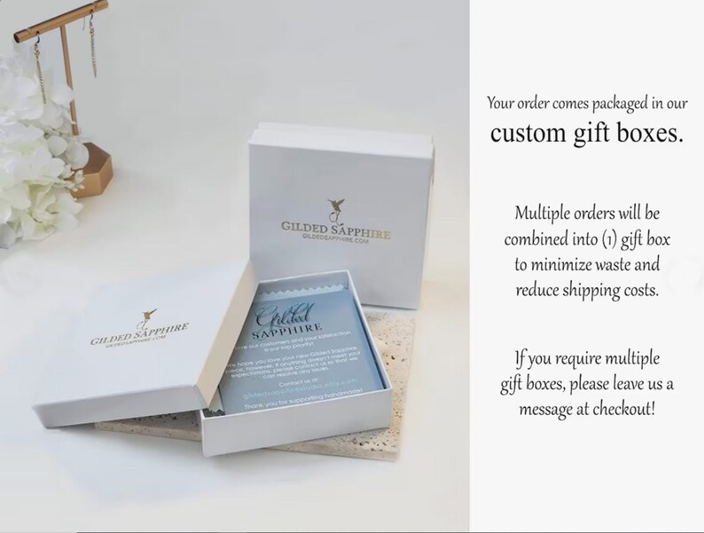 K&ouml;nnte beinhalten: Wei&szlig;e Geschenkbox mit einem Kolibri-Logo und dem Text "Gilded Sapphire" auf der Oberseite. Die Box ist ge&ouml;ffnet und eine kleinere wei&szlig;e Box mit einer blauen Karte darin ist sichtbar. Die Karte tr&auml;gt den Text "Gilded Sapphire". Der Text "Ihre Bestellung wird in unseren individuellen Geschenkboxen verpackt." ist auf der rechten Seite des Bildes zu sehen.
