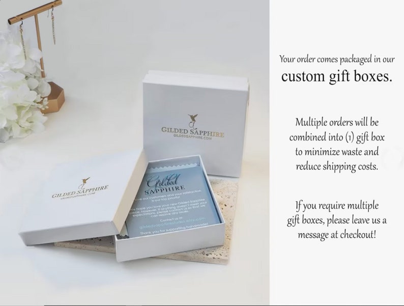 May include: A white square cardboard gift box with a gold Gilded Sapphire logo, featuring a matching lid and a light blue informational card inside. The text on the box reads Gilded Sapphire, and the card includes contact information for the brand.