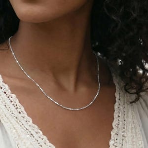 Puede incluir: Un delicado collar de plata con pequeñas cuentas redondas y reflectantes. El collar se lleva sobre una blusa de encaje blanco. La imagen es un primer plano, centrado en el collar y la parte superior del pecho.
