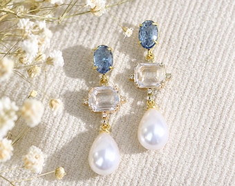 Op de afbeelding: Een paar goudkleurige oorbellen met een blauwe ovale kristal aan de bovenkant, een rechthoekige heldere kristal in het midden en een witte druppelvormige parel aan de onderkant, verbonden door kleine gouden schakels en kleine steentjes.