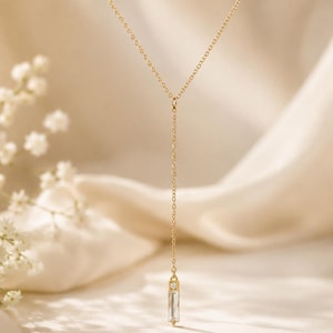 May include: A gold-toned Y-shaped necklace featuring a delicate chain and a vertical pendant. The pendant consists of a small, clear, rectangular baguette-cut stone topped with a tiny, round, clear accent stone, creating a minimalist drop design.